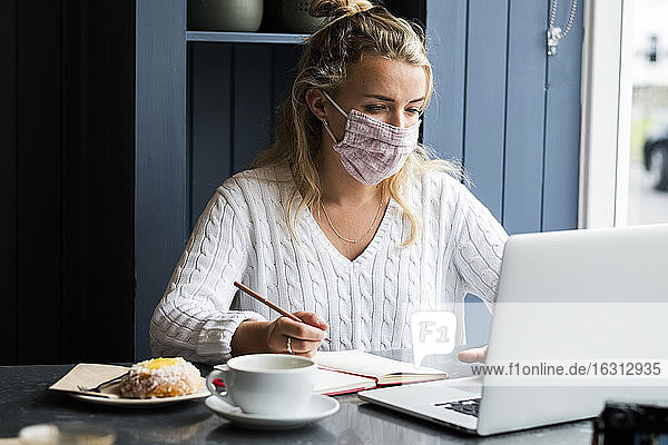 Frau mit Gesichtsmaske  die allein an einem Café-Tisch mit einem Laptop-Computer sitzt und aus der Ferne arbeitet.