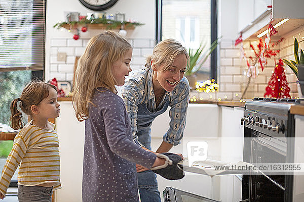 Blonde Frau mit blauer Schürze und zwei Mädchen stehen in der Küche und backen Weihnachtsplätzchen.
