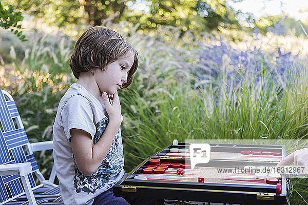 Ein kleiner Junge spielt Backgammon im Freien in einem Garten.