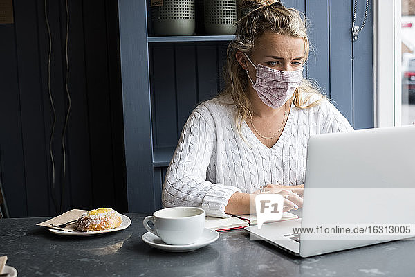 Frau mit Gesichtsmaske  die allein mit einem Laptop an einem Cafétisch sitzt und aus der Ferne arbeitet.