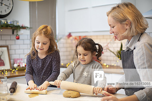 Blonde Frau mit blauer Schürze und zwei Mädchen stehen in der Küche und backen Weihnachtsplätzchen.