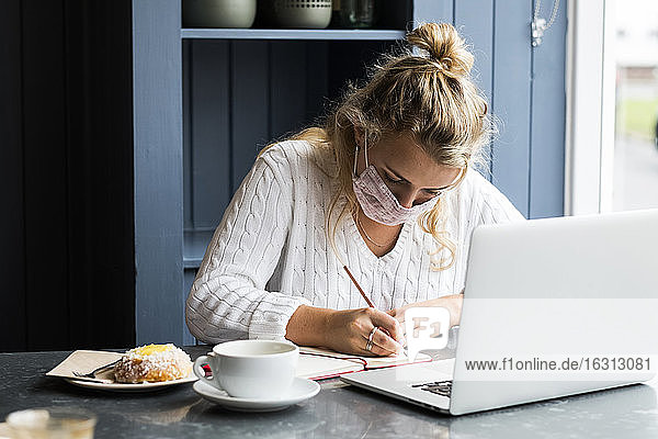 Junge blonde Frau mit Gesichtsmaske sitzt allein mit einem Laptop-Computer an einem Café-Tisch