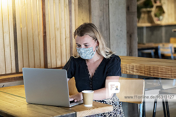 Young blond woman wearing blue face mask  sitting at table  using laptop computer.