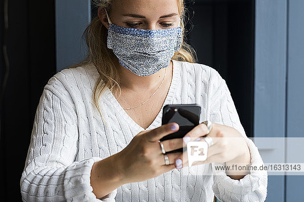 Junge blonde Frau mit Gesichtsmaske  die allein in einem Café sitzt  ein Mobiltelefon benutzt und aus der Ferne arbeitet.