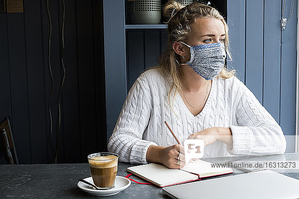 Frau mit Gesichtsmaske  die allein an einem Café-Tisch sitzt und einen Laptop benutzt  der aus der Ferne arbeitet.