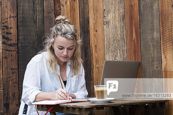 Young blond woman sitting alone at a cafe table with a laptop working remotely.