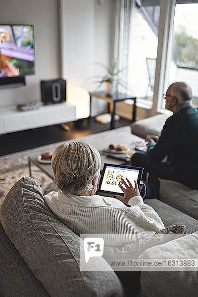 Hochwinkelansicht einer älteren Frau mit digitalem Tablet  während der männliche Partner auf dem Sofa im Wohnzimmer sitzt