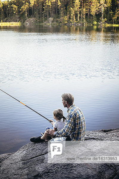 Father and daughter fishing while sitting by lake