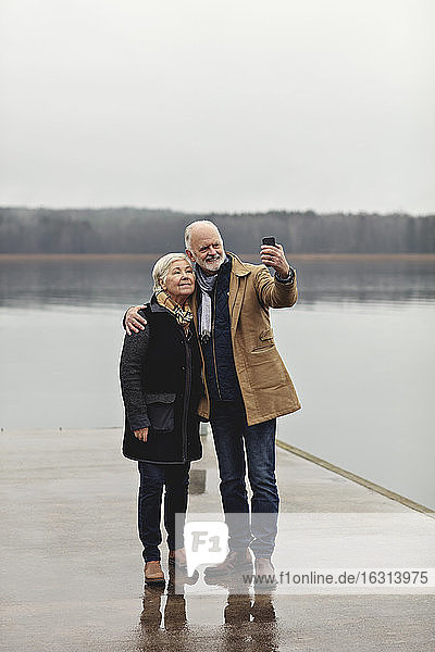 Full length of senior couple taking selfie at lakeside against clear sky