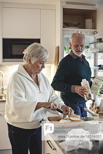 Senior woman preparing food while male partner holding drink in kitchen