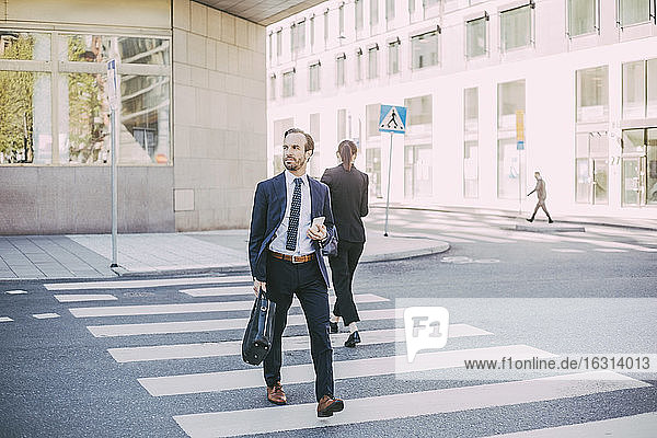 Businessman with phone looking away while crossing road in city