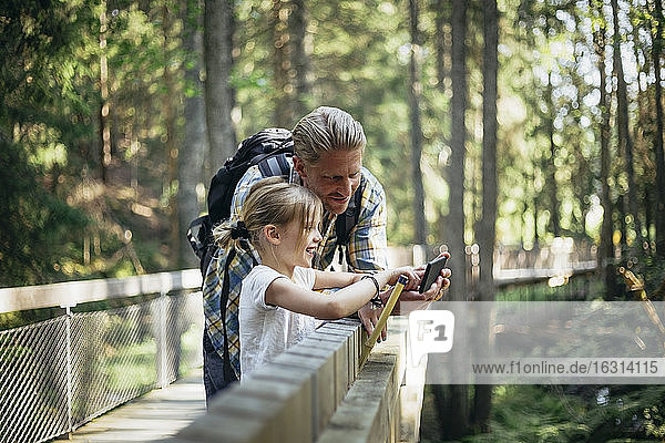 Lächelnder Vater mit Rucksack und Tochter beim Blick auf Smartphone im Wald