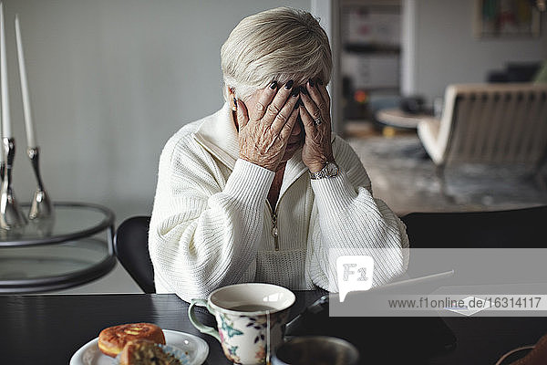 Besorgte ältere Frau mit dem Kopf in der Hand  die zu Hause am Esstisch sitzt