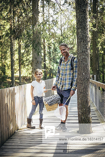 Portrait of father and daughter with picnic basket standing on footbridge in forest