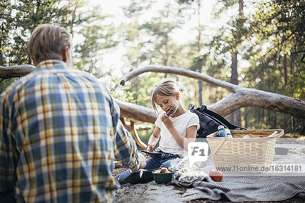 Daughter eating watermelon while sitting with father on picnic blanket in forest