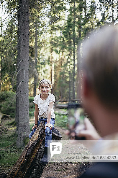 Lächelnde Tochter sitzt auf einem Baumstamm  während der Vater im Wald mit einem Smartphone fotografiert