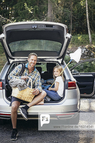 Porträt eines lächelnden Vaters mit Picknickkorb der im Kofferraum sitzenden Tochter