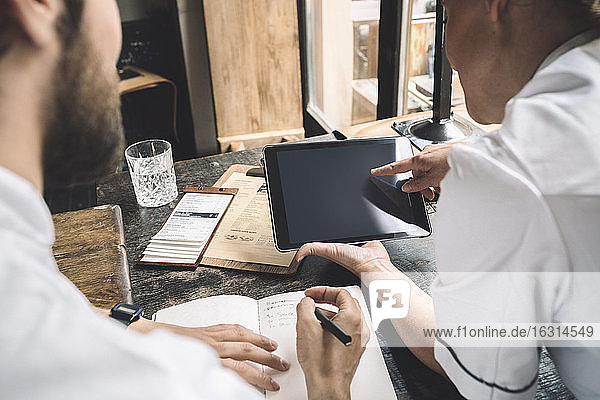 Midsection of chef with digital tablet while coworker writing at table in restaurant