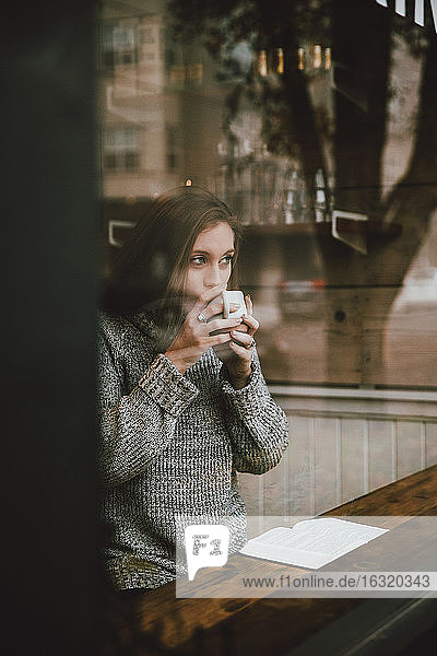 Young woman drinking coffee and reading book at cafe window
