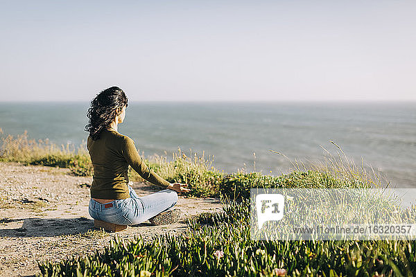 Gelassene Frau meditiert auf einer sonnigen Klippe mit Blick auf das Meer