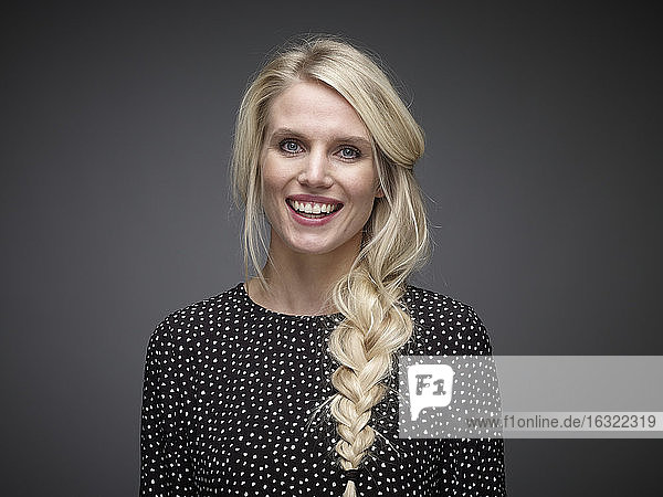 Porträt einer lächelnden blonden jungen Frau mit Zopf vor einem grauen Hintergrund