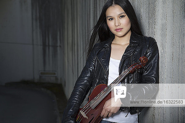 Porträt einer jungen Asiatin mit Geige  die an einer Betonwand lehnt