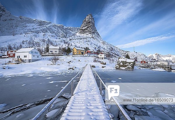 Fußgängerbrücke über zugefrorenen Fjord  hinten Wohnhäuser in verschneiter Landschaft und Bergformation Hammarskaftet  Nordland  Lofoten  Norwegen  Europa