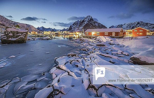 Eisschollen am zugefrorenem Fjord  hinten rote Holzhäuser und winterliche Berglandschaft in der Abenddämmerung  Reine  Nordland  Lofoten  Norwegen  Europa