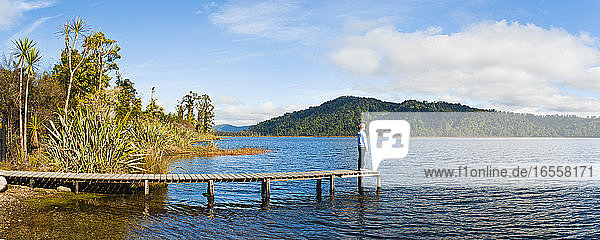 Panoramafoto eines Touristen  der die Ruhe am Lake Ianthe genießt  Westküste  Südinsel  Neuseeland. Dieses Panoramafoto wurde am Lake Ianthe aufgenommen  einem der zahlreichen Höhepunkte der Fahrt entlang der Westküste der neuseeländischen Südinsel.