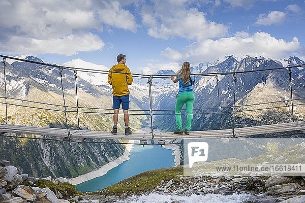 Wanderer  junge Frau und junger Mann auf Hängebrücke an der Olpererhütte  Ausblick auf Schlegeisstausee  Speicher Schlegeis  hinten Zillertaler Alpen mit Gletscher Schlegeiskees  Zillertal  Tirol  Österreich  Europa