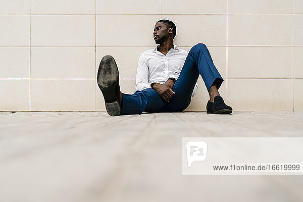 Müder afrikanischer Unternehmer  der wegschaut  während er auf dem Fußweg an der Wand sitzt und sich entspannt