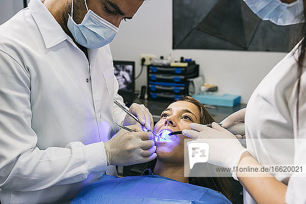 Männlicher Zahnarzt in Maske und Handschuhen untersucht die Zähne einer Patientin mit Hilfe einer Assistentin in einer Klinik