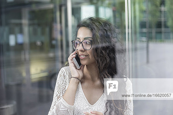 Nahaufnahme einer Frau mit langen Haaren  die am Fenster stehend über ein Mobiltelefon spricht