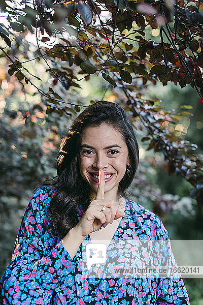 Nahaufnahme einer lächelnden jungen Frau mit Finger auf den Lippen  die im Park vor Pflanzen steht