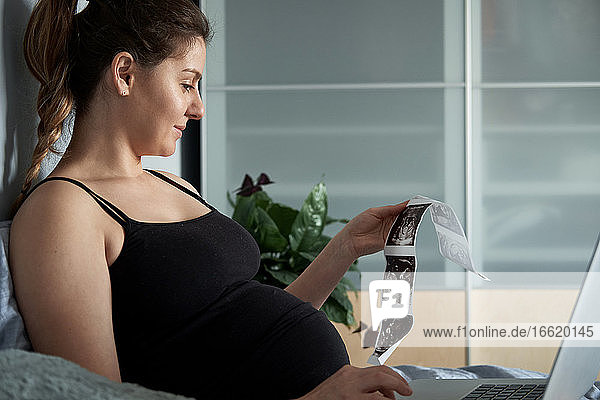 Schwangere Frau hält Ultraschall-Scans  während sie einen Laptop auf dem Bett zu Hause benutzt
