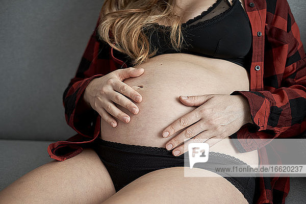Nahaufnahme einer schwangeren Frau mit Händen auf dem Bauch  die zu Hause auf dem Sofa sitzt