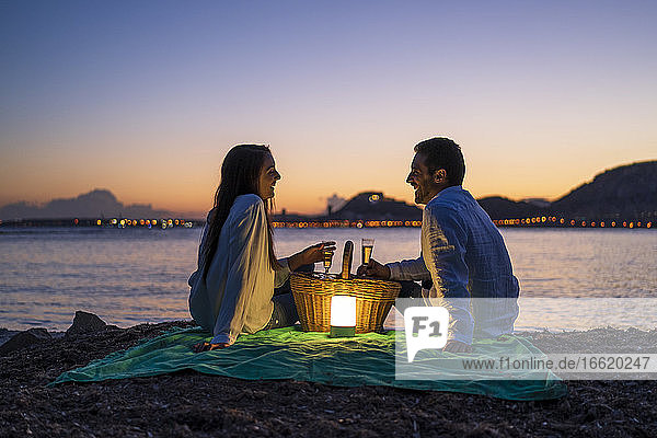 Couple with drinks looking at each other while sitting on beach against clear sky during dusk