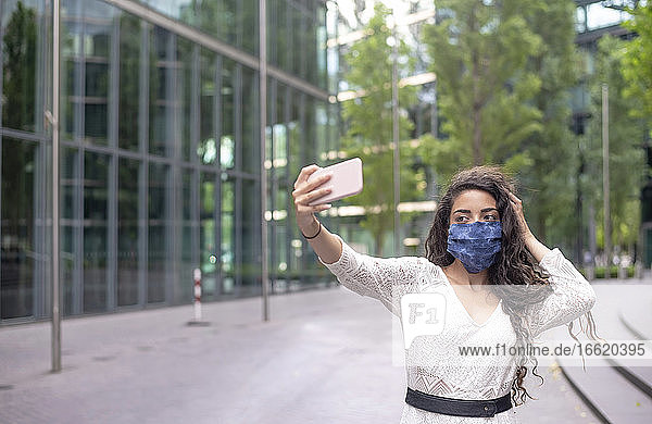 Junge Frau mit Maske  die ein Selfie mit ihrem Smartphone macht  während sie auf einem Fußweg in der Stadt steht