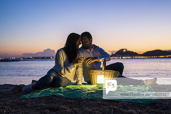 Romantisches Paar mit Getränken am Strand sitzend gegen den klaren Himmel in der Abenddämmerung