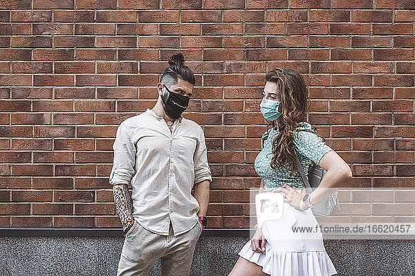 Couple in protective face masks standing against brick wall during coronavirus outbreak