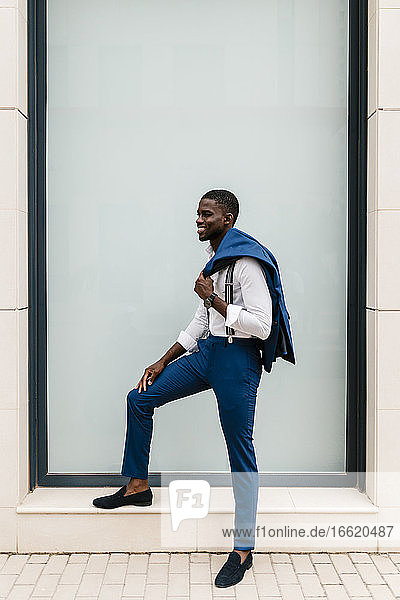 Lächelnder junger männlicher Unternehmer  der einen Anzug hält und am Fenster in der Stadt steht