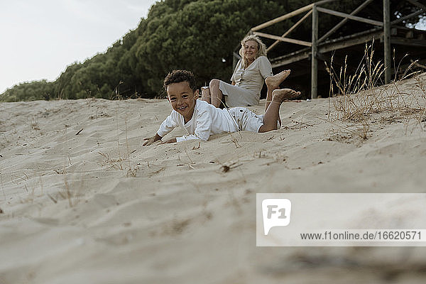Grandmother sitting while grandson lying on sand at beach