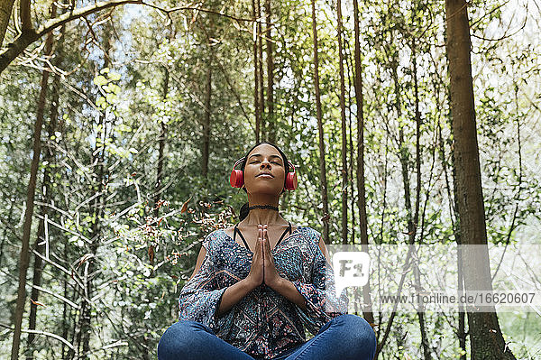 Young woman with eyes closed listening music through headphones while meditating against trees at park