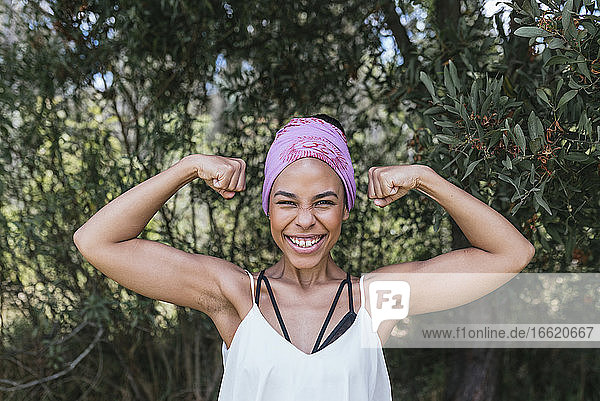 Glückliche Frau mit lila Kopftuch  die ihre Muskeln anspannt  während sie im Park vor Pflanzen steht
