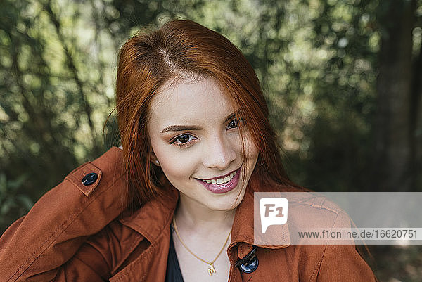 Lächelnde schöne junge rothaarige Frau mit brauner Jacke im Park
