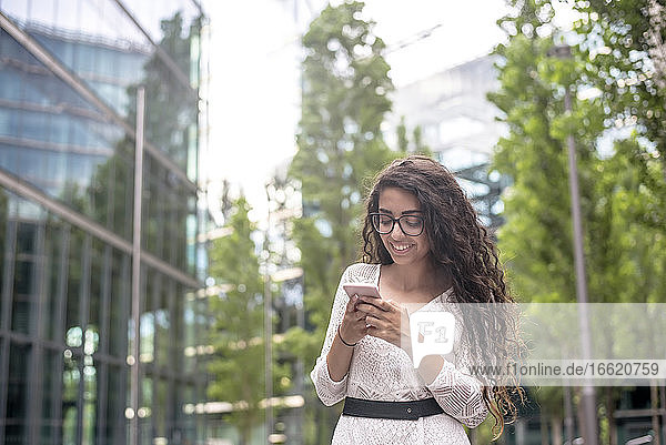 Lächelnde junge Frau mit langen Haaren  die ein Mobiltelefon benutzt  während sie in der Stadt steht