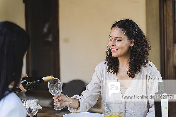 Woman pouring champagne in friend's wineglass while sitting by table at back yard