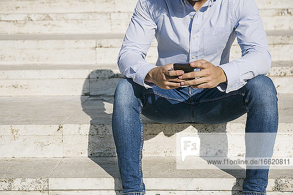 Mann surft im Internet über sein Smartphone  während er an einem sonnigen Tag auf einer Treppe sitzt