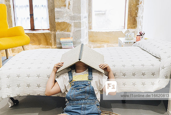 Kleines Mädchen bedeckt ihr Gesicht mit einem Buch  während sie zu Hause im Schlafzimmer sitzt