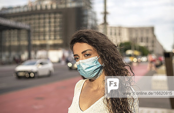 Junge Frau mit Maske  die wegschaut  während sie auf einem Fußweg in der Stadt steht
